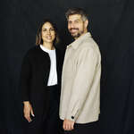 Studio portrait of married couple Wassmer against a dark backdrop: a woman in a black coat with a white top and a man in a light beige overshirt, both smiling at the camera.