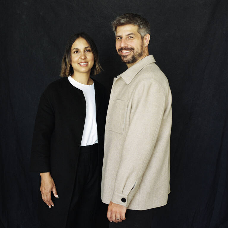 Studio portrait of married couple Wassmer against a dark backdrop: a woman in a black coat with a white top and a man in a light beige overshirt, both smiling at the camera.