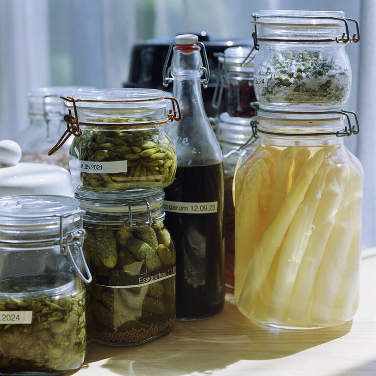 Assortment of preserved ingredients in jars: white asparagus in brine, pickles, buds, and a dark bottle (e.g., garum), labeled and sunlit.