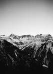 Black-and-white photo of a mountain range with snow-covered peaks beneath a clear sky; dark forested slopes in the foreground.